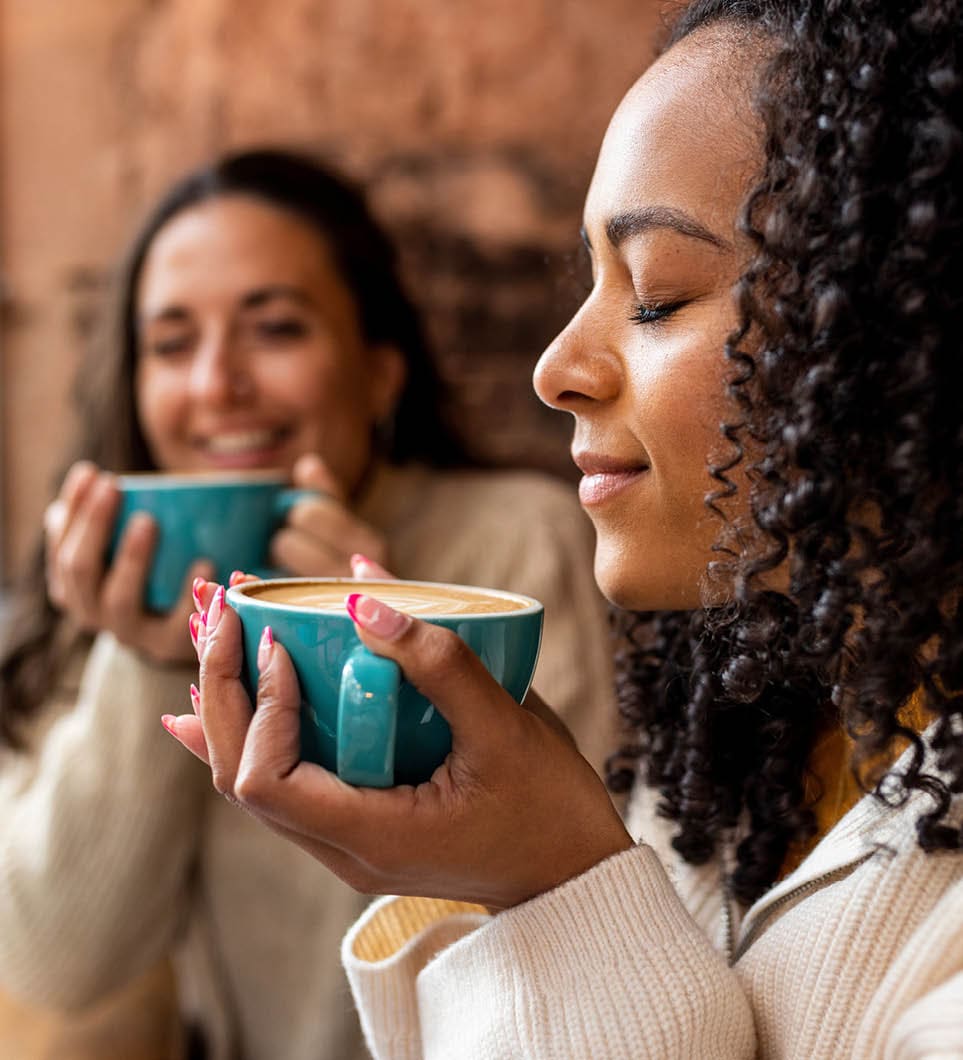 Woman with curly hair smelling coffee by friend in background at cafe