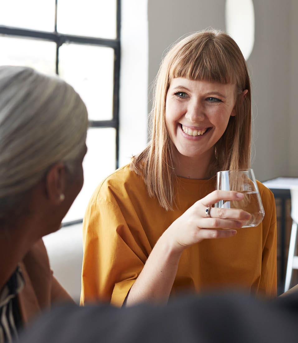 Smiling businesswoman with drinking glass looking at female coworker while sitting in modern workplace
